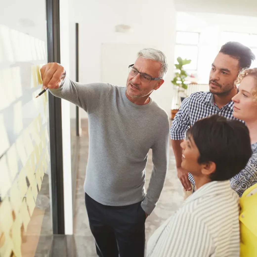 businesspeople-brainstorming-with-adhesive-notes-in-a-meeting-1180532869 Mature businessman and his diverse team brainstorming with yellow adhesive notes on a glass wall in a bright modern office