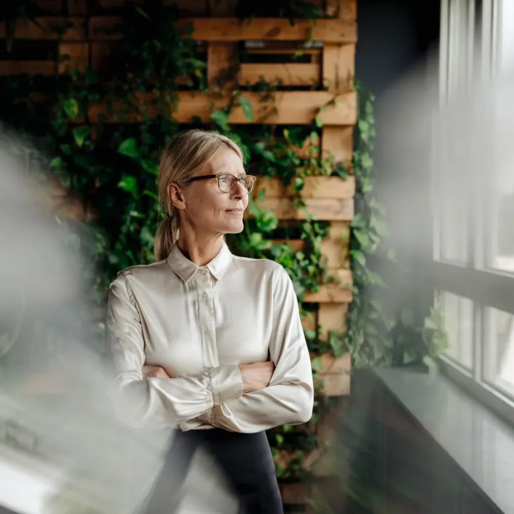 businesswoman-in-green-office-looking-out-of-window--1056320180 Eine Frau mit Brille steht am Fenster und betrachtet die Außenwelt. Sie trägt eine weiße Bluse und hat die Arme in stiller Zuversicht verschränkt. Das Grün und die Holzvertäfelung hinter ihr spiegeln die Agilität wider, die sie verkörpert – ähnlich wie agile Produktionsplanung in dynamischen Umgebungen.