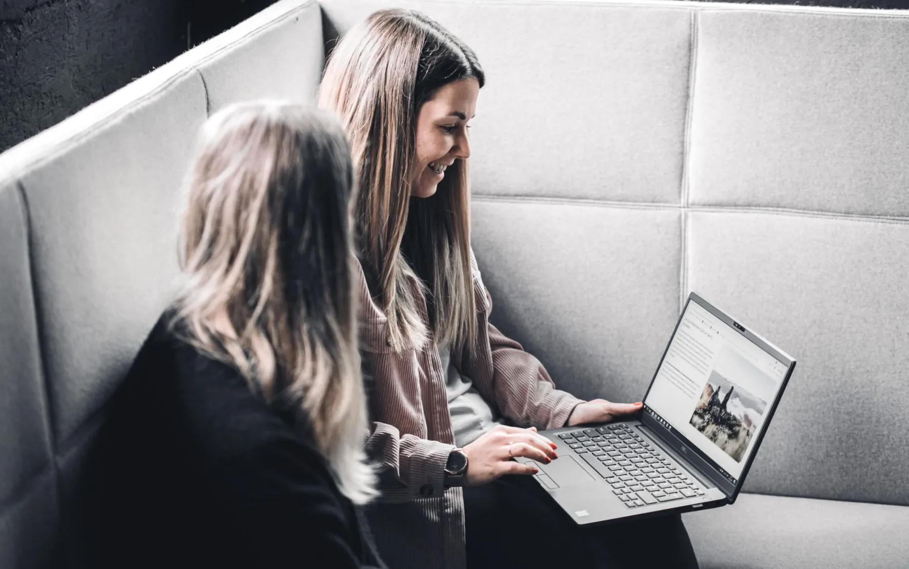 Digitalisierung im Tourismus ist unsere Leidenschaft: valantic austria Zwei Frauen sitzen auf einem grauen Sofa; eine zeigt der anderen auf einem Laptop Reiseinhalte, die die neuesten Fortschritte in der Reisetechnologie vorstellen.
