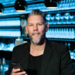 A man with a beard and short hair holds a smartphone in a dimly lit bar with rows of illuminated bottles in the background.