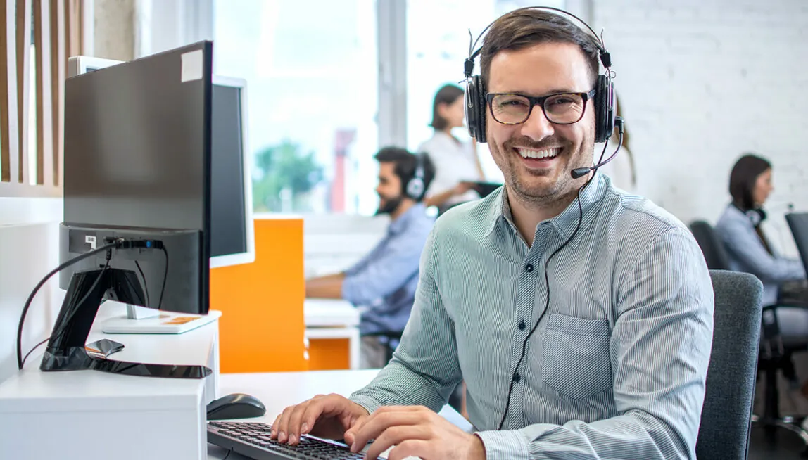 Mann sitzt mit Headset am Schreibtisch und schaut lachend in die Kamera Mann sitzt mit Headset am Schreibtisch und schaut lachend in die Kamera