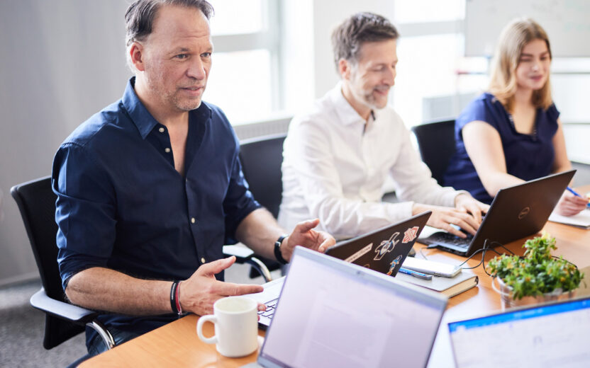 Drei Personen sitzen mit Laptops an einem Konferenztisch und diskutieren. Auf dem Tisch stehen Laptops, eine Kaffeetasse und eine Pflanze. Ein Mann spricht, während der andere Mann und die andere Frau an ihren Laptops arbeiten.