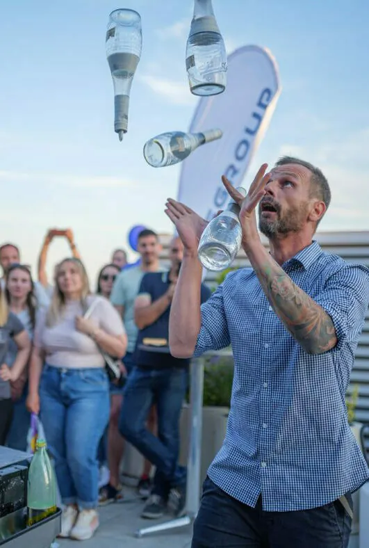 Ein Mann mit einer Tätowierung auf dem Unterarm jongliert im Freien mit vier durchsichtigen Flaschen, während im Hintergrund eine Menschenmenge zusieht.