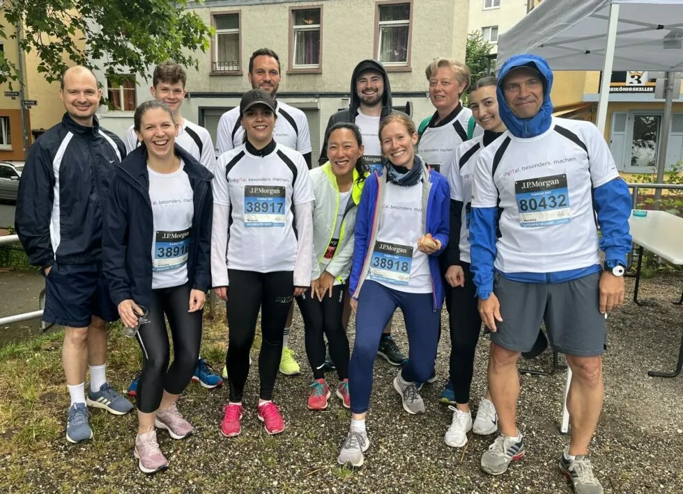 Eine Gruppe von Menschen in Sportkleidung versammelt sich vor einem Rennen im Freien. Jeder trägt ein passendes weißes Hemd und eine Startnummer. Im Hintergrund sind Wohnhäuser und ein Registrierungstisch zu sehen.