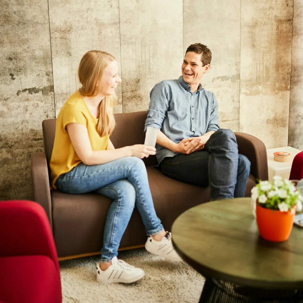 Zwei Personen sitzen auf einer Couch und unterhalten sich. Sie lächeln sich in einem warm beleuchteten Raum an. Im Vordergrund steht ein runder Tisch mit einer Topfpflanze.