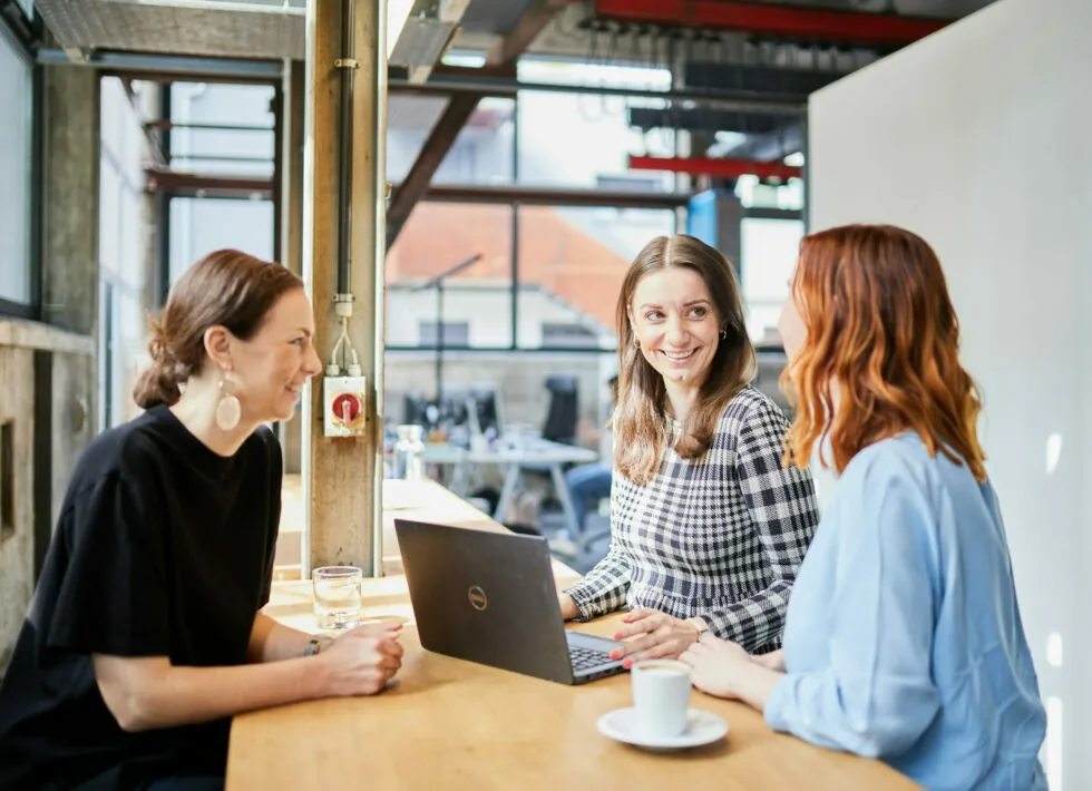 Drei Frauen diskutieren an einem Holztisch mit einem Laptop und Getränken in einem modernen, gut beleuchteten Büroumfeld.