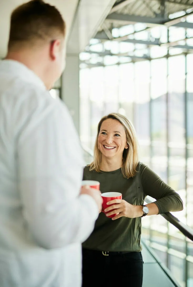 Zwei Personen unterhalten sich und halten rote Kaffeetassen in einem hellen, modernen Innenraum mit großen Fenstern.