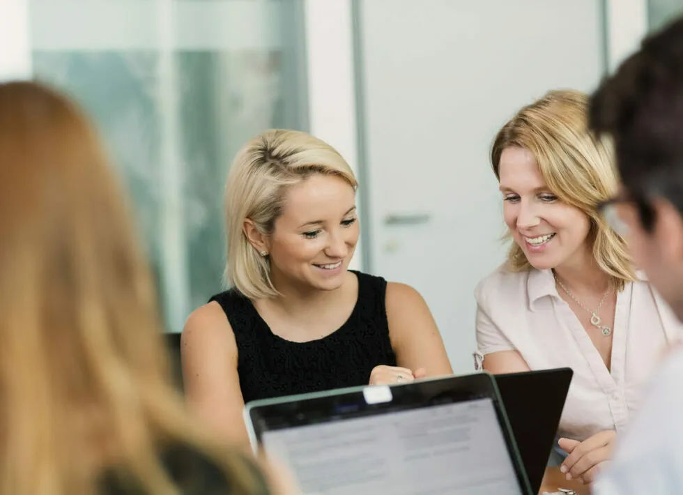 Zwei Frauen sitzen an einem Tisch, schauen auf einen Laptop und lächeln. Um sie herum sind teilweise andere Personen zu sehen.