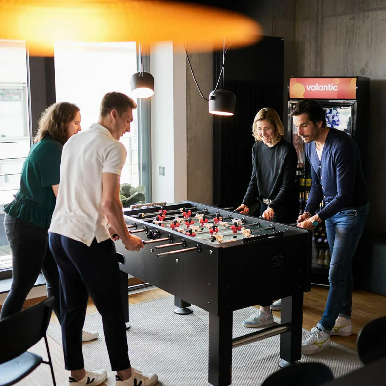Vier Personen sitzen um einen Tischfußballtisch herum und spielen in einem gut beleuchteten, modernen Raum aktiv ein Spiel. Im Hintergrund ist ein Verkaufsautomat zu sehen.