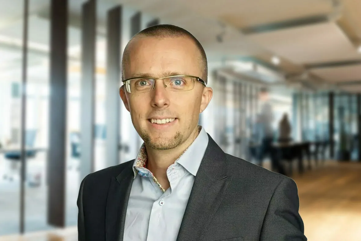 Ein Mann mit Brille und Anzugjacke über einem Hemd mit Knöpfen steht in einem modernen Büro mit Glaswänden und Holzböden im Hintergrund.