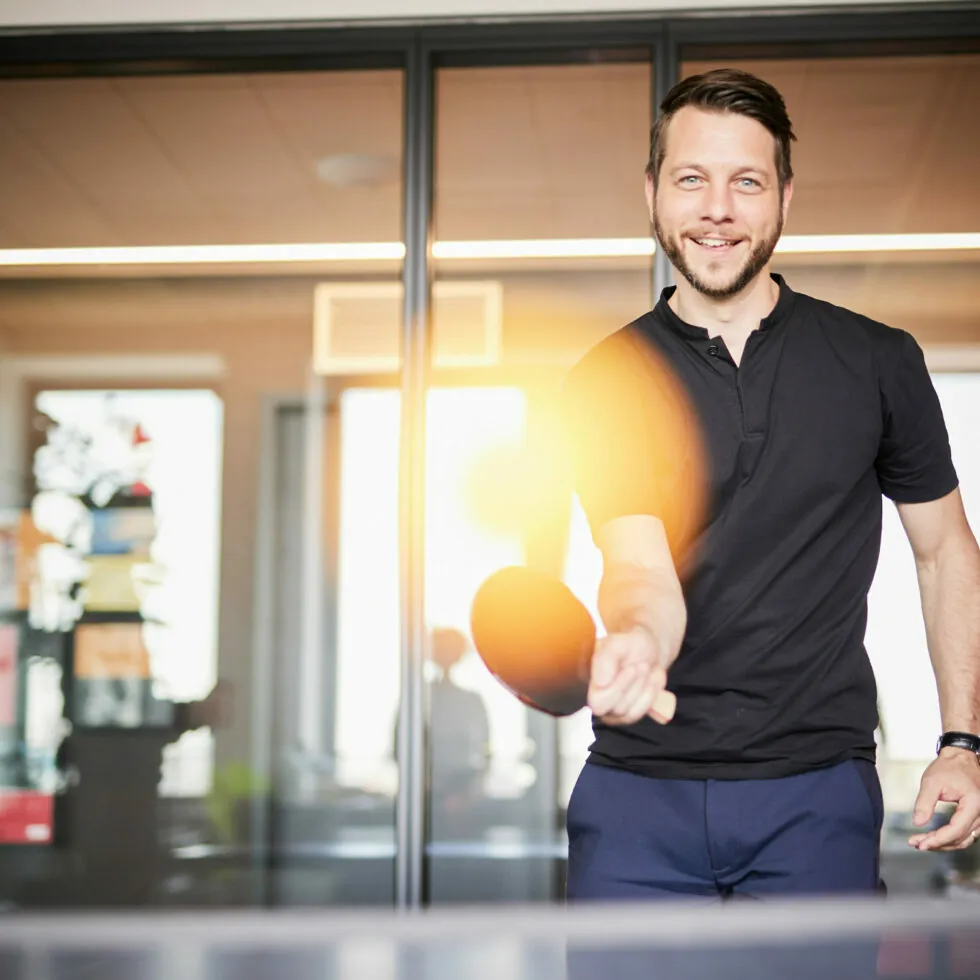 Ein Mann im schwarzen Hemd lächelt, während er drinnen Tischtennis spielt, während neben seinem Schläger helles Licht aufflackert.