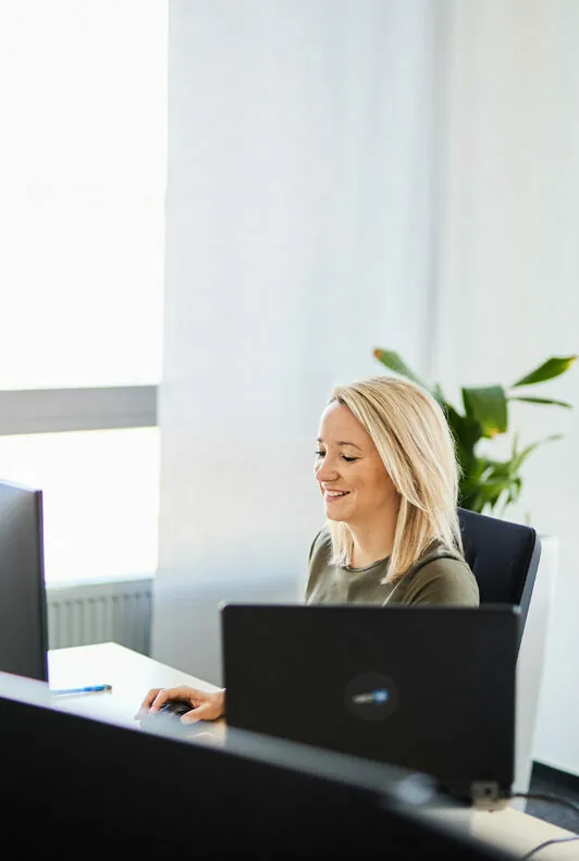 Eine Frau mit blonden Haaren arbeitet in einem hellen Büro an einem Computer, im Hintergrund steht eine Pflanze.
