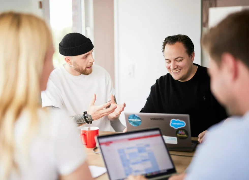 Zwei Männer unterhalten sich, einer trägt eine Beanie und gestikuliert, während der andere lächelt und an einem Laptop arbeitet. Im Vordergrund sind außerdem eine verschwommene Frau und eine weitere Person zu sehen, die wegblickt.