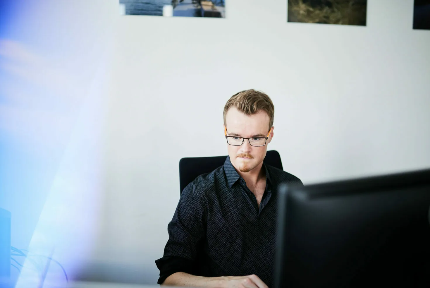 Ein Mann mit Brille und schwarzem Hemd sitzt konzentriert an einem Schreibtisch und arbeitet am Computer.