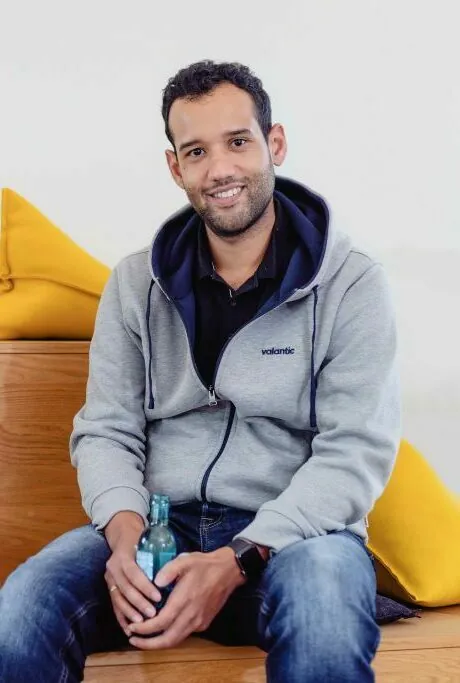 Ein Mann in grauem Kapuzenpullover und Jeans sitzt auf einer Holzbank und hält eine Flasche in der Hand. Im Hintergrund sind gelbe Kissen zu sehen. Er lächelt und blickt in die Kamera.