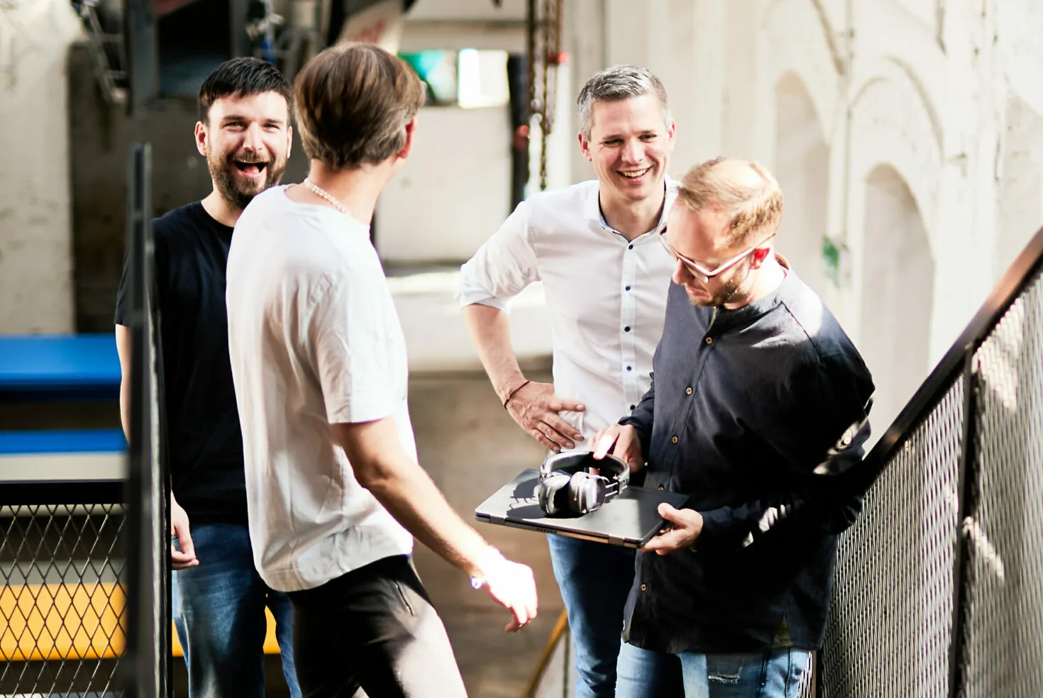 Vier Männer stehen auf einer Treppe und unterhalten sich. Einer hält einen Laptop und Kopfhörer in der Hand, die anderen lächeln und hören zu. Sie befinden sich in einem hellen Raum im Industriestil.