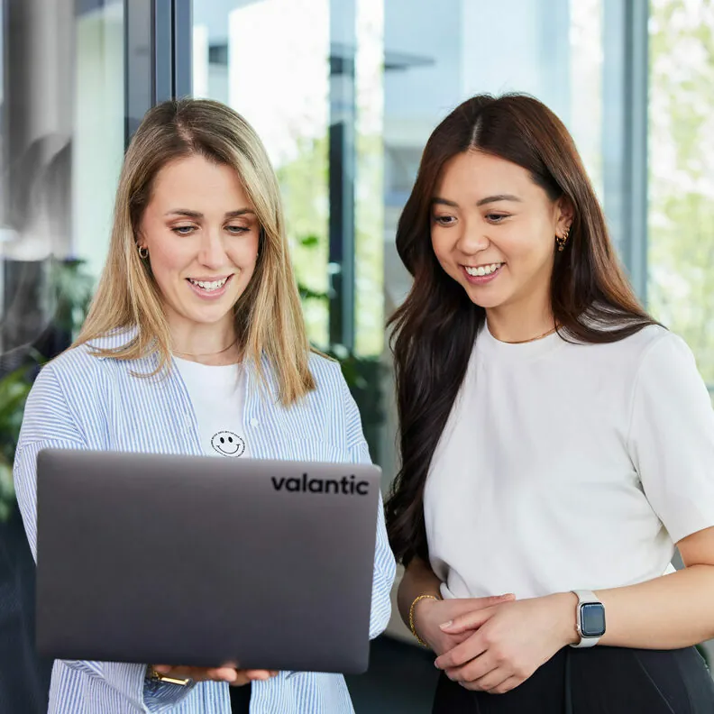 Zwei Frauen stehen drinnen, lächeln und schauen auf einen Laptop mit dem Logo „Valantic“ auf der Rückseite. Eine Frau hat blondes Haar und die andere langes dunkles Haar.