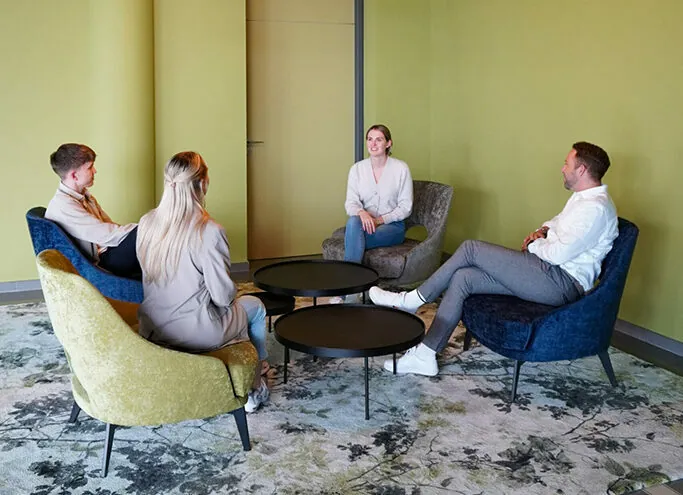 In einem modernen Büro sitzen vier Personen im Halbkreis auf Loungesesseln und unterhalten sich an drei runden Couchtischen.