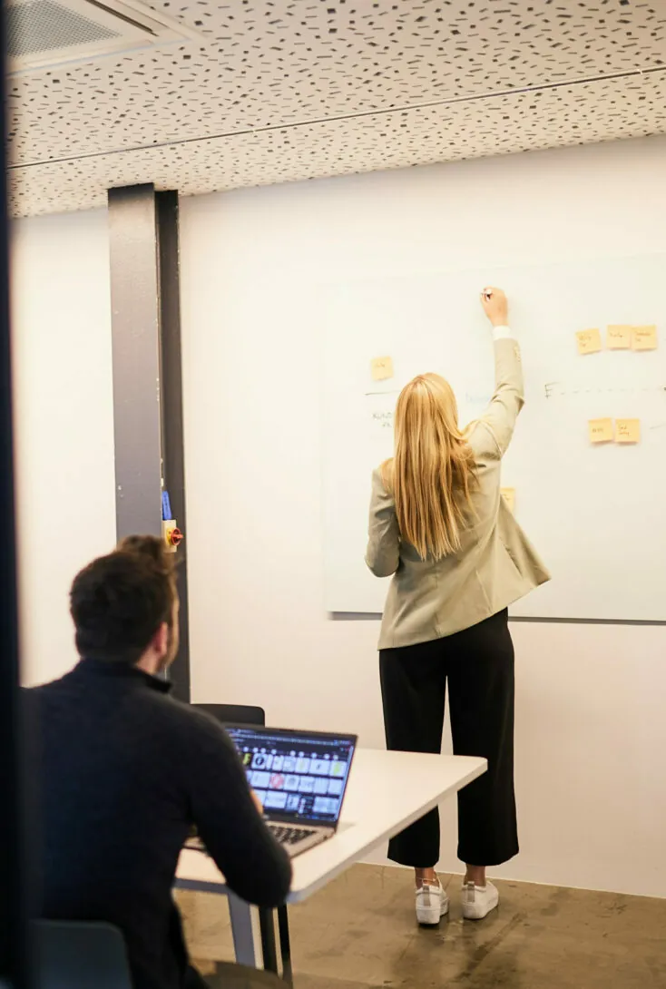 Eine Frau schreibt mit Haftnotizen auf ein Whiteboard, während ihr ein Mann mit einem geöffneten Laptop an einem Tisch gegenübersitzt.