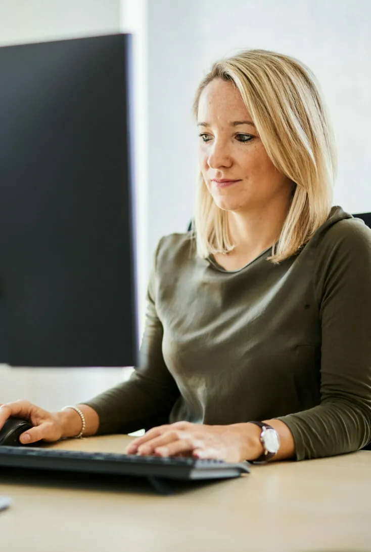 Eine Frau mit schulterlangen blonden Haaren und einem grünen Oberteil arbeitet in einem Büro an einem Computer. Sie schaut auf den Monitor und tippt auf einer Tastatur.