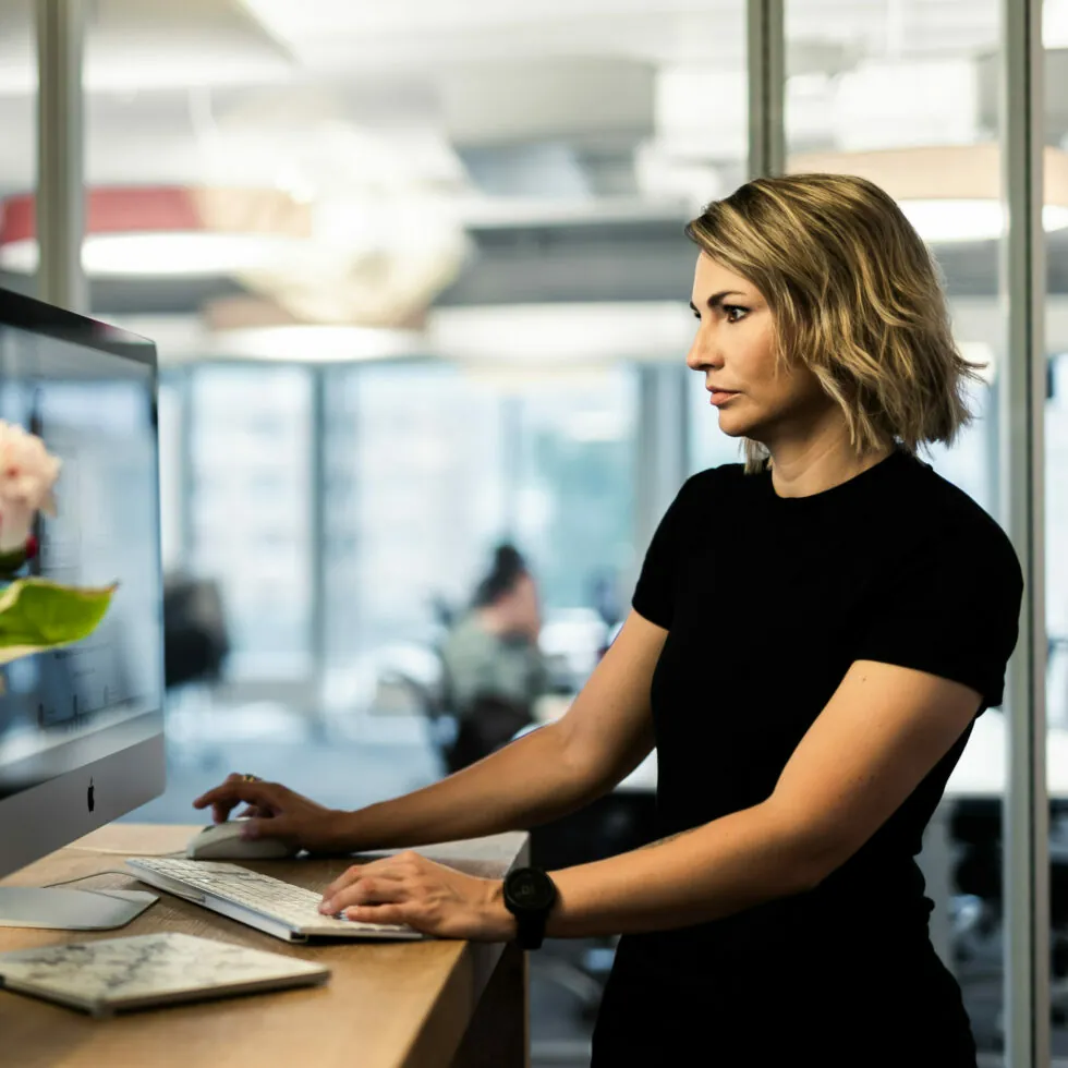 Eine Person mit kurzen Haaren steht an einem Schreibtisch und benutzt einen Computer in einer modernen Büroumgebung. Im Hintergrund sind andere Personen zu sehen, die in einem offenen Raum arbeiten.