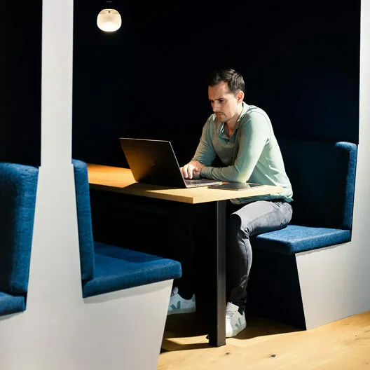 Eine Person sitzt allein in einer schwach beleuchteten Kabine und arbeitet an einem Laptop. Die Kabine verfügt über einen Holztisch und gepolsterte Sitze.