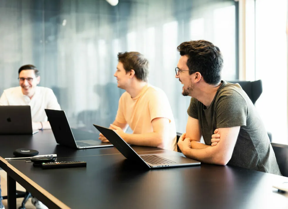 Drei Männer sitzen an einem Tisch in einem Büro und unterhalten sich. Jeder von ihnen hat einen Laptop vor sich. Licht aus großen Fenstern erhellt den Raum.