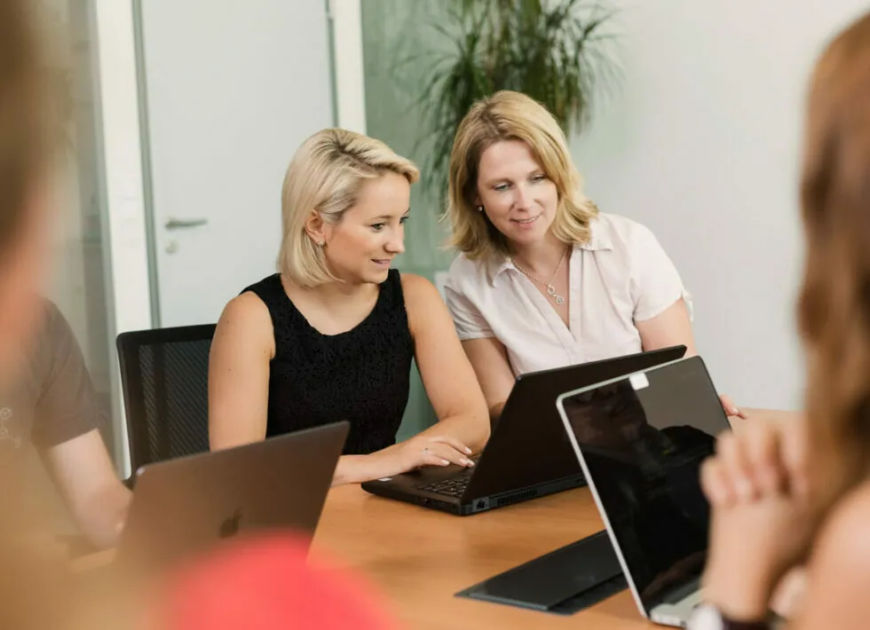 Zwei Frauen sitzen an einem Konferenztisch und arbeiten gemeinsam an einem Laptop. Vor ihnen stehen weitere geöffnete Laptops und im Vordergrund sind unscharfe Menschen zu sehen.