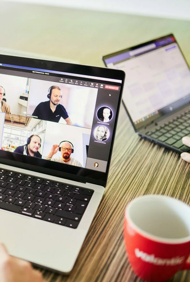 Auf einem Laptop wird ein Gruppen-Videoanruf mit fünf Teilnehmern angezeigt. Ein weiterer Laptop und eine rote Tasse stehen auf dem Holztisch.