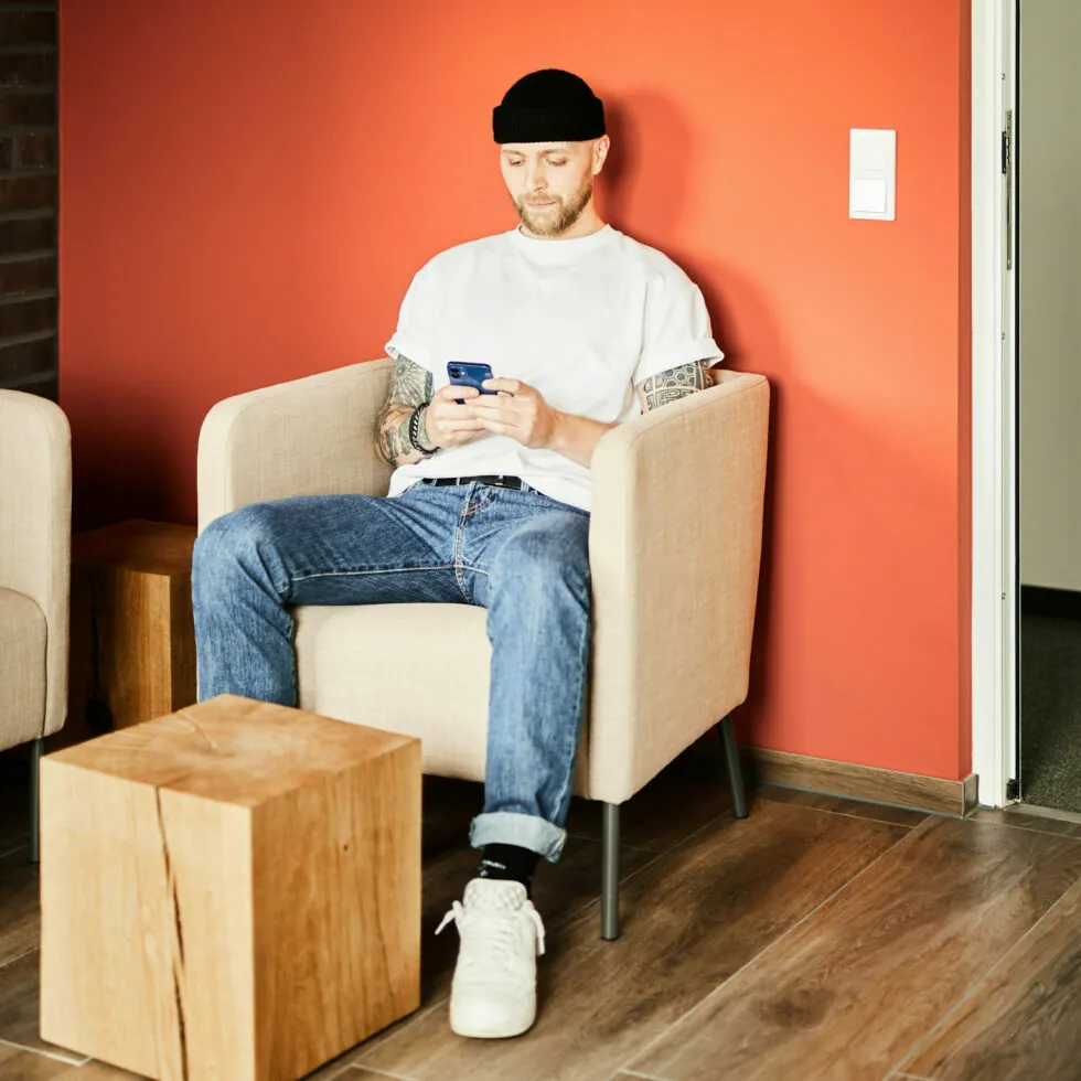 Ein Mann mit schwarzer Beanie-Mütze und weißem T-Shirt sitzt in einem beigen Sessel und telefoniert in einem modernen Raum mit einer roten Wand und einem hölzernen Beistelltisch.