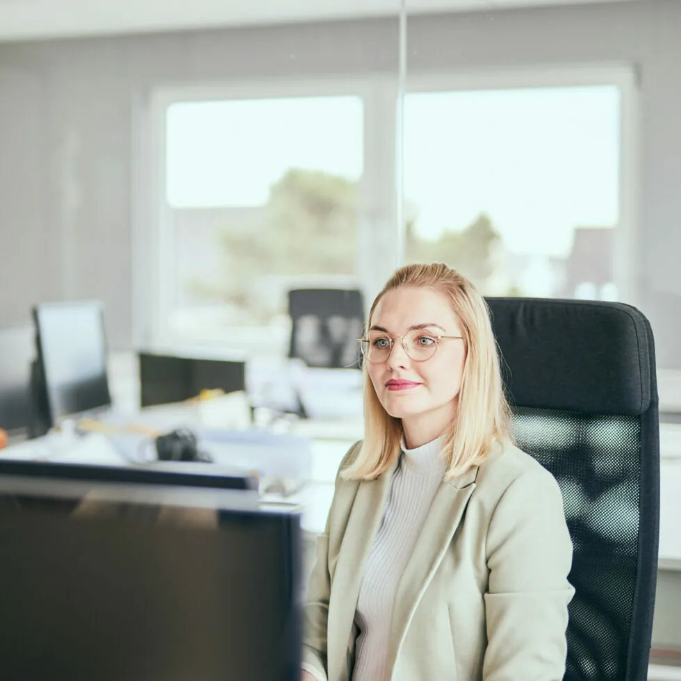 Eine Person mit Brille und hellem Blazer sitzt an einem Schreibtisch in einem Büro und schaut auf einen Computerbildschirm. Das Büro verfügt über mehrere Schreibtische, Computer und im Hintergrund ein großes Fenster.