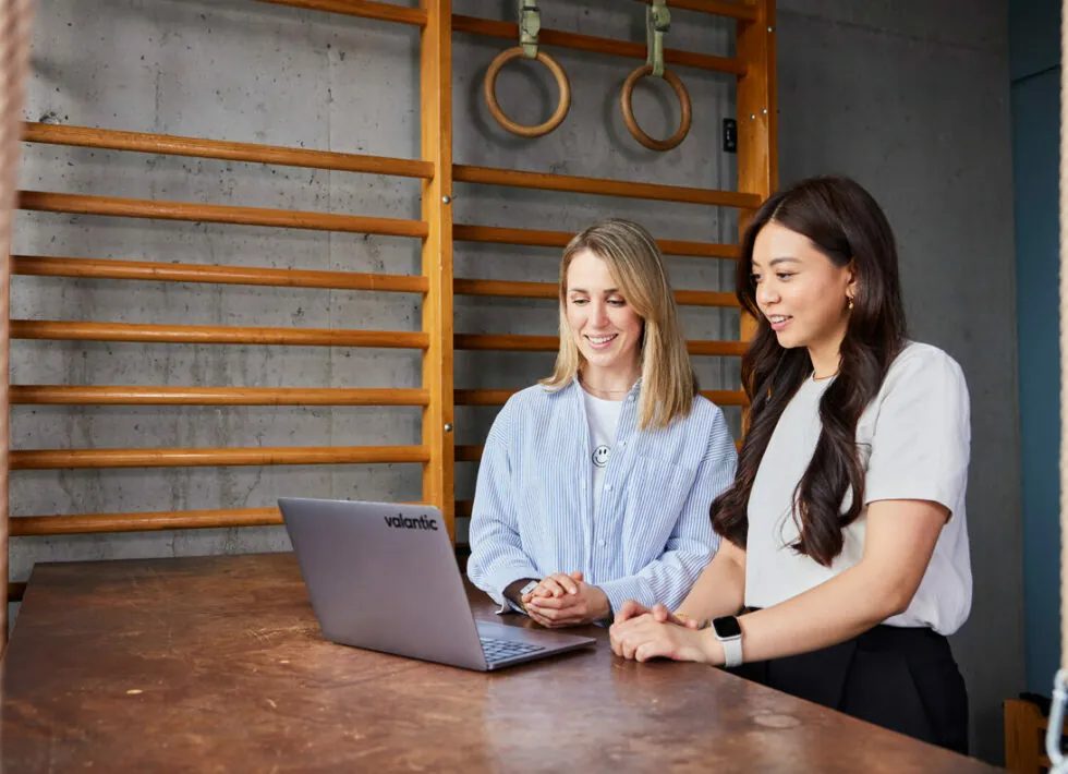 Zwei Frauen stehen an einem Holztisch, beide lächeln und schauen auf einen Laptop-Bildschirm. Im Hintergrund sind Turnringe und ein hölzernes Sprossenwand-Reck an einer Wand zu sehen.