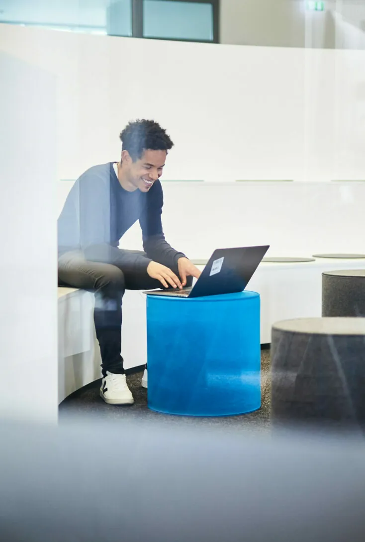 Eine Person in dunklem Pullover und Hose sitzt auf einer Bank und benutzt einen Laptop, der auf einem blauen Hocker steht. Die Person lächelt und schaut auf den Laptop-Bildschirm.