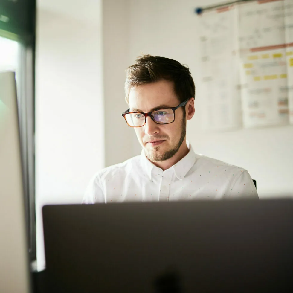 Ein Mann mit Brille sitzt in einem gut beleuchteten Raum mit einem Whiteboard und einem Kalender im Hintergrund und schaut auf einen Laptop-Bildschirm.