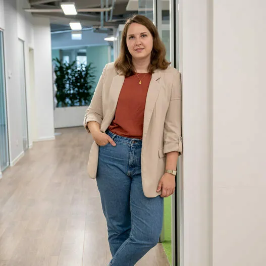Eine Frau in beigem Blazer und Jeans steht lässig an einer Wand in einem modernen Büroflur. Sie hat eine Hand in der Tasche und blickt in die Kamera.