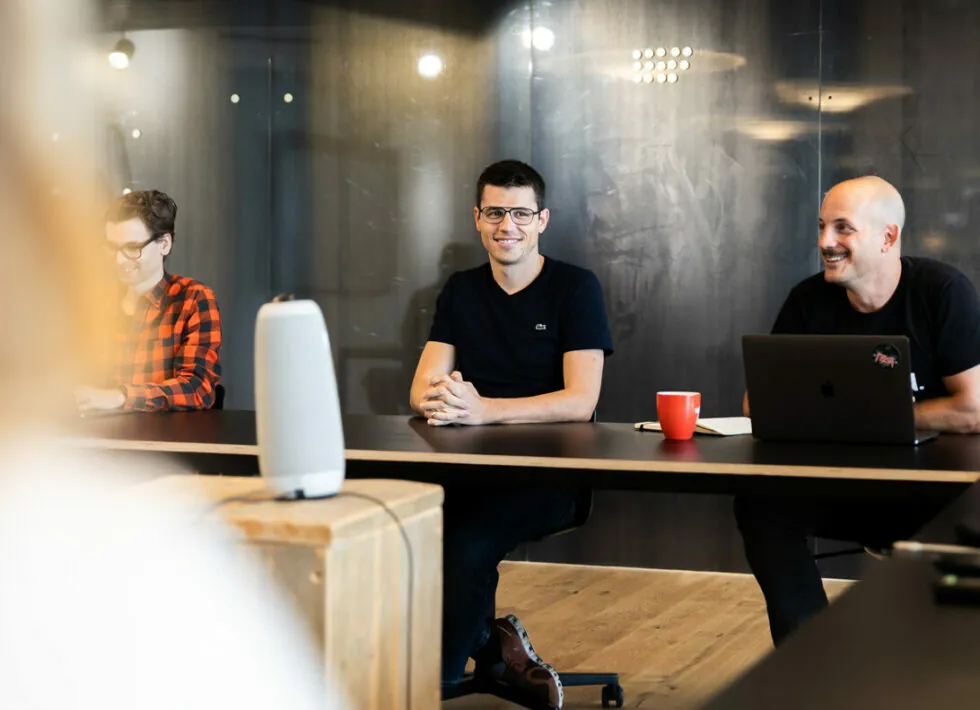 Drei Personen in einem Besprechungsraum sitzen an einem Tisch mit einem Laptop, einer Kaffeetasse und einem intelligenten Lautsprecher. Eine Person ist im Fokus und lächelt in die Kamera, während die anderen beiden in die Diskussion vertieft sind.