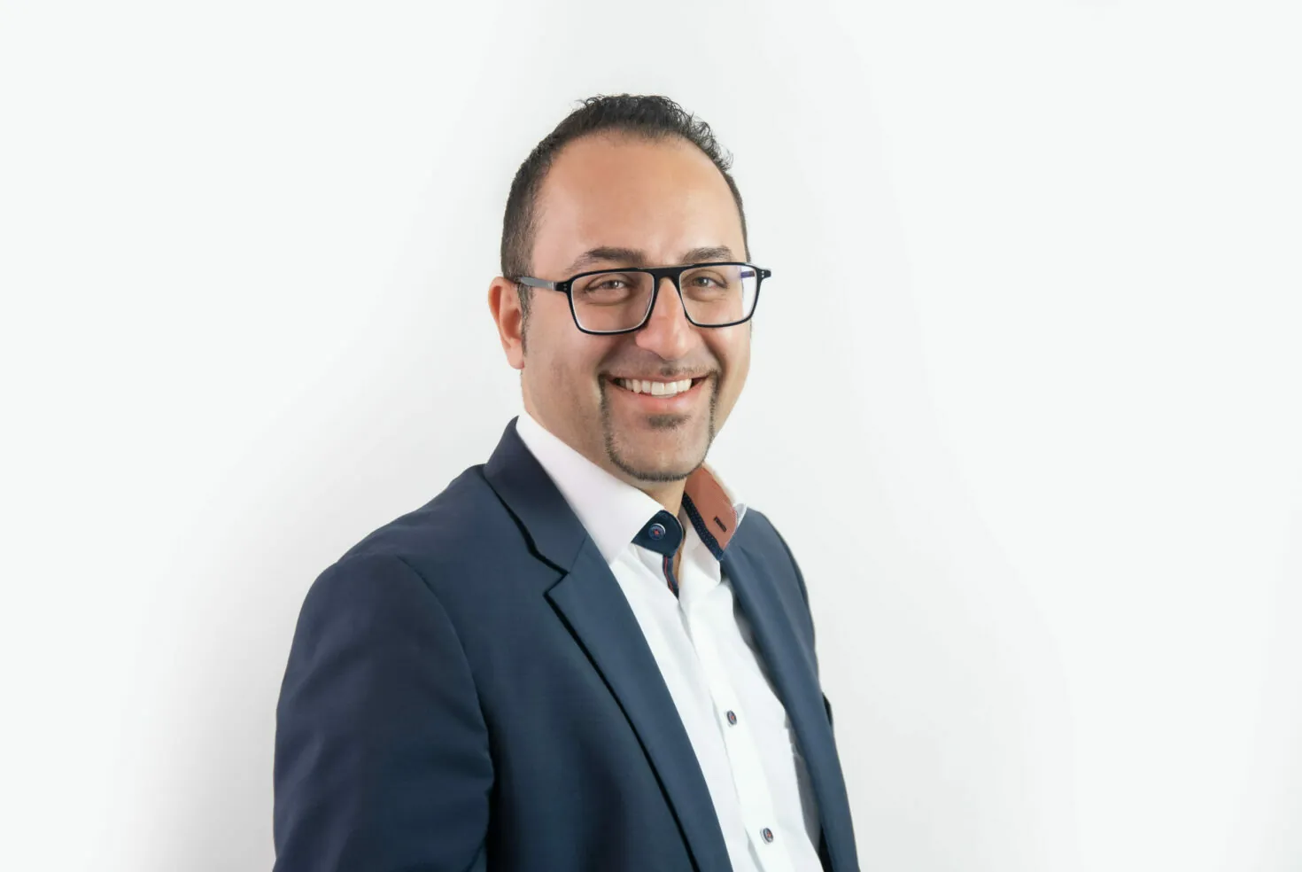 Lächelnder Mann mit Brille und kurzen dunklen Haaren, bekleidet mit weißem Hemd und dunklem Blazer, vor einem schlichten weißen Hintergrund.