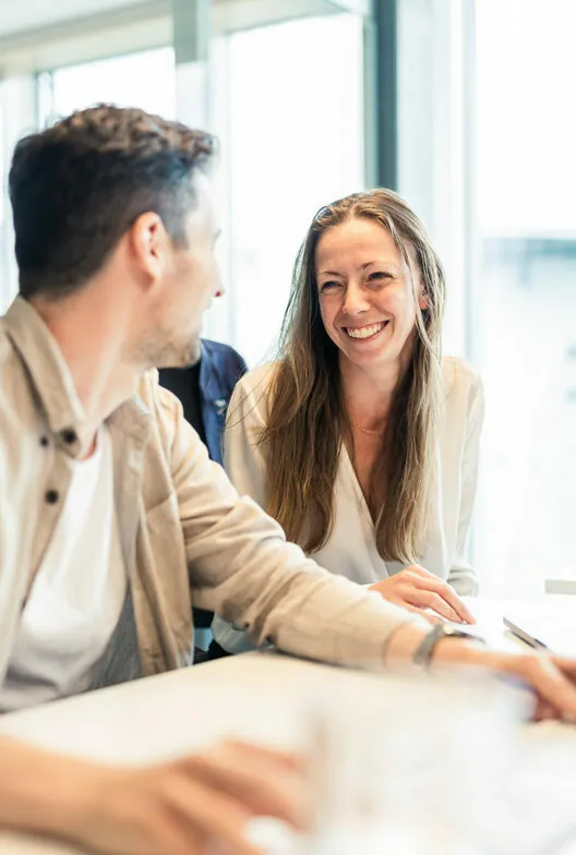 Zwei Personen sitzen drinnen an einem Tisch und unterhalten sich. Die Person rechts lächelt die Person links an. Durch die Fenster im Hintergrund fällt helles Tageslicht.