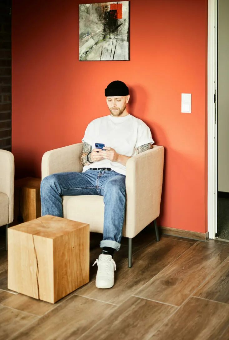 Ein Mann mit Bart und Tattoos sitzt auf einem beigen Sessel vor einer roten Wand und schaut auf sein Handy. Er trägt ein weißes T-Shirt, Jeans und eine schwarze Beanie-Mütze. In der Nähe stehen ein Holzwürfel und ein Gemälde.