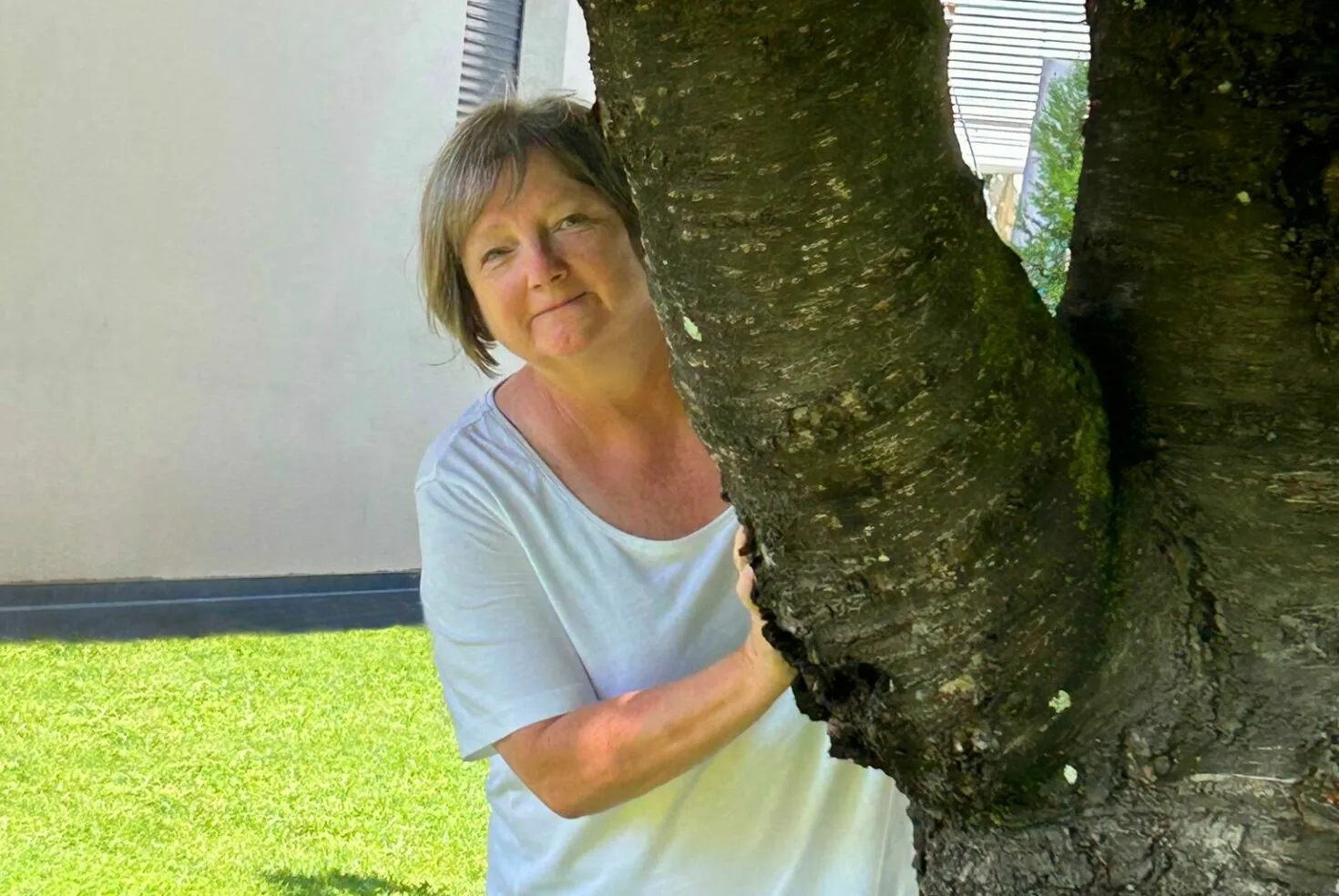 Eine Frau mit kurzen Haaren und weißem Hemd steht teilweise versteckt hinter einem Baumstamm und lächelt in die Kamera.
