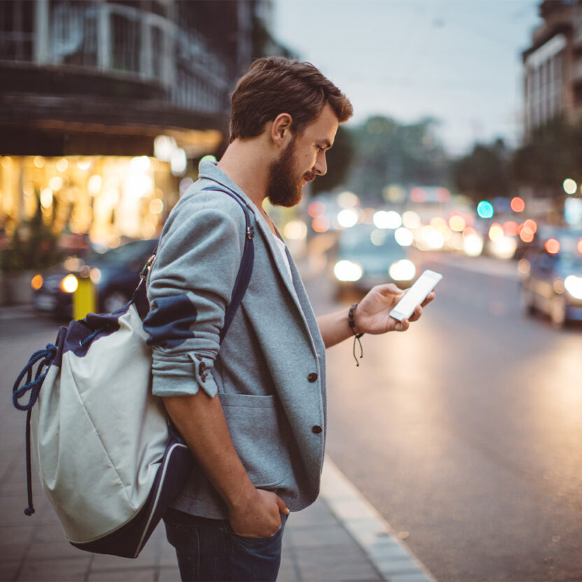 young-man-on-the-streets-of-big-city-495457462 Young man on the street of big city , waiting taxi, chacking his smartphone, for news or new messages. Or looking for map instructions. Carry backpack on one shoulder. Dusk time. Casual dressed. Street lights in background.