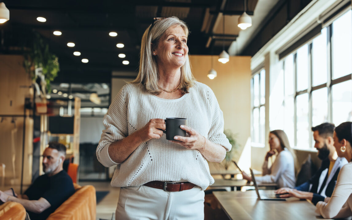 businesswoman-looking-away-thoughtfully-1354221337 Thoughtful businesswoman standing in a co-working space. Mature businesswoman smiling cheerfully while holding a cup of coffee. Experienced entrepreneur contemplating new ideas during her coffee break.