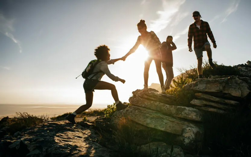 Group of hikers on a mountain. Woman helping her friend to climb a rock. Young people on mountain hike at sunset.