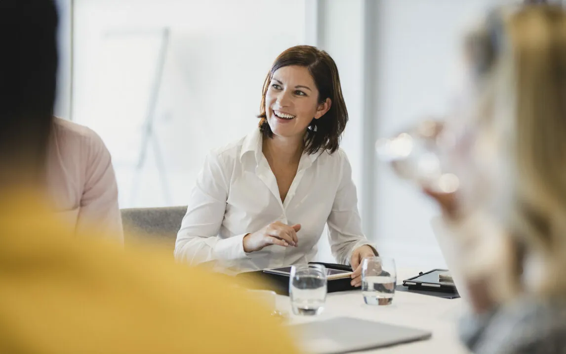 woman-smiling-at-business-meeting-1200927481 Businesswoman smiling at meeting table, listening, learning, success, happiness
