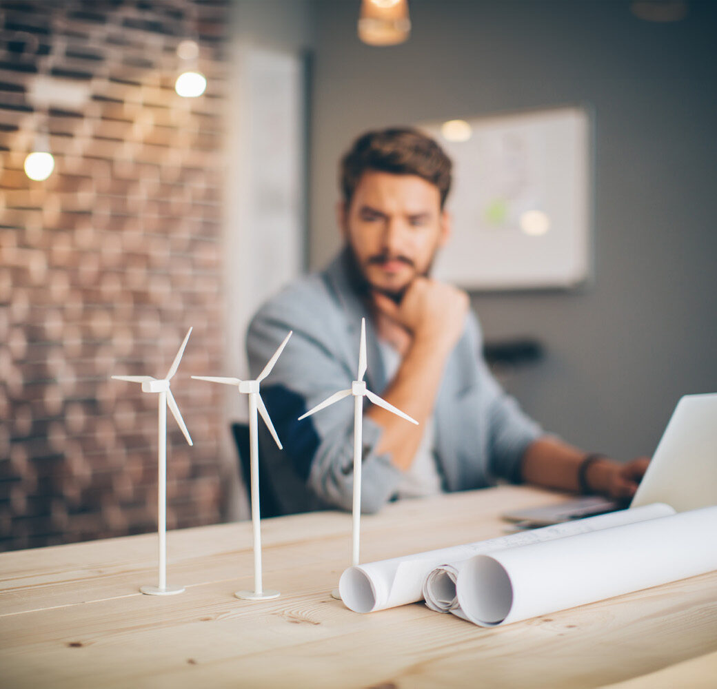 Young engineer at modern office space, sitting at the table and working on energy project. Focus on wind turbine models on the table.
