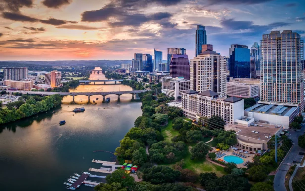 Aerial view of a city skyline with tall buildings, a river with bridges, and green spaces at sunset.