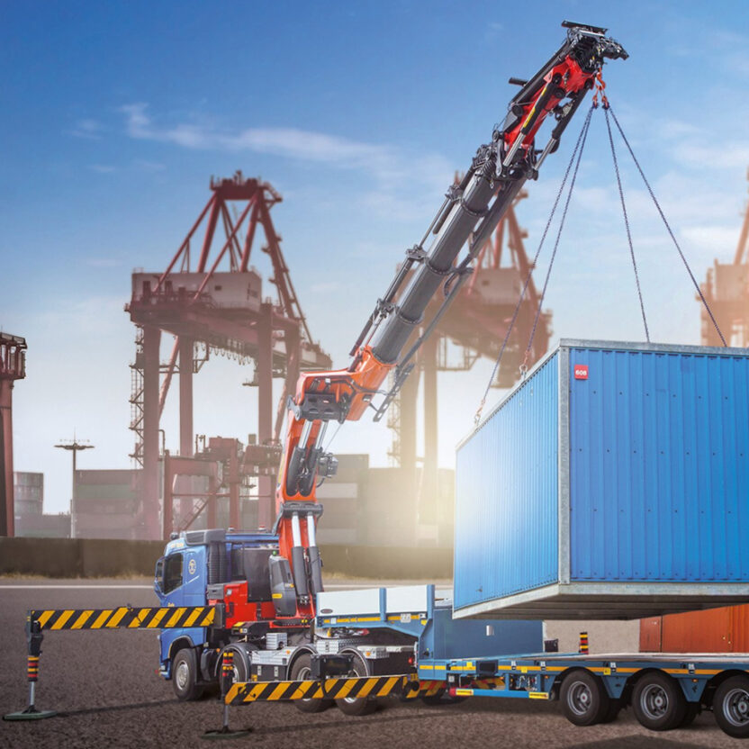 Ein Kranwagen hebt in einem großen Verladehof einen blauen Frachtcontainer auf einen Anhänger. Im Hintergrund sind Stapel bunter Container und Frachtkräne unter einem klaren Himmel zu sehen.