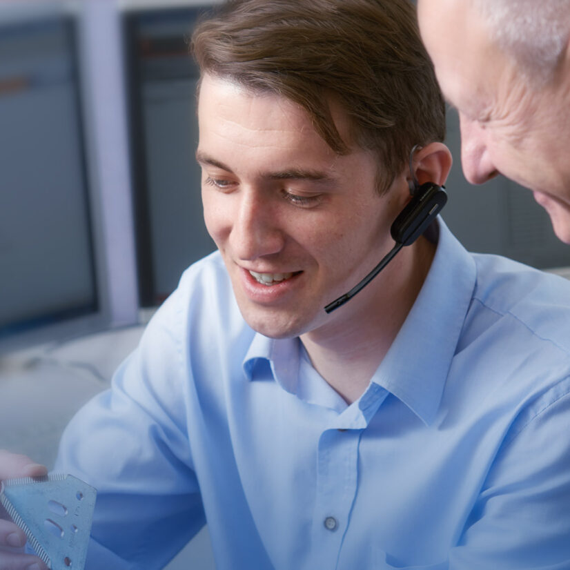 Zwei Männer in einem Büro untersuchen einen kleinen dreieckigen Gegenstand. Einer trägt ein Headset und lächelt, der andere schaut interessiert zu. Im Hintergrund sind Computer zu sehen.