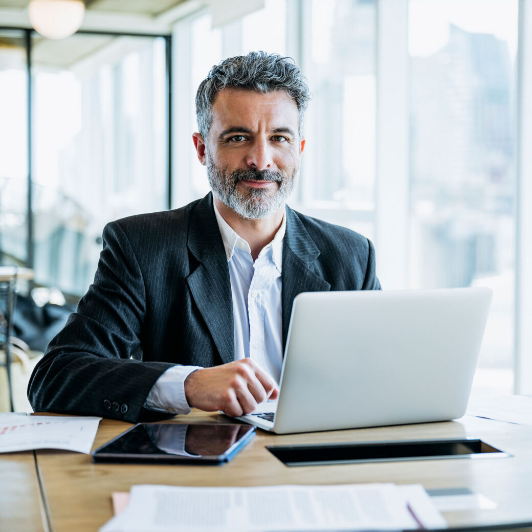 Businessman sitting at office work table next to window with view of downtown and pausing from typing on laptop to smile at camera.
