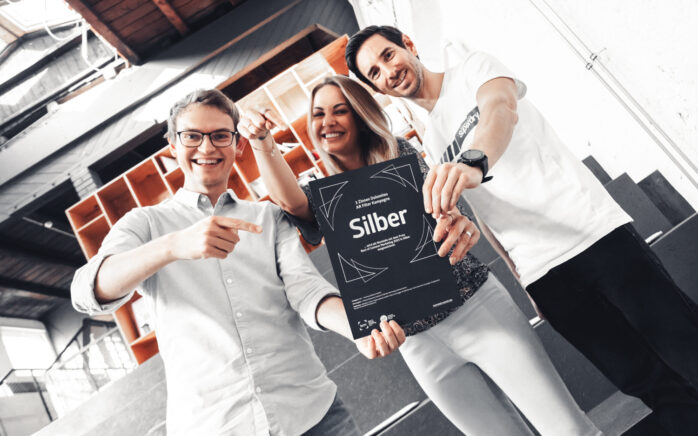Eine Frau und zwei Männer halten freudestrahlend eine Silber-Auszeichnung in ihren Händen.
